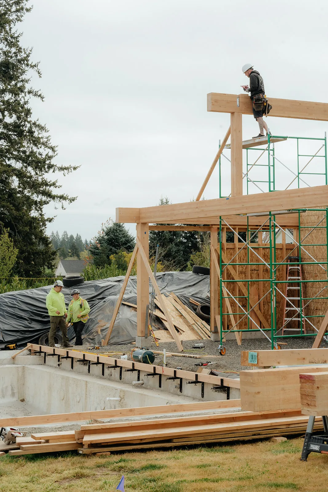 Timber pavilion frame under construction with scaffold and crew in a residential yard